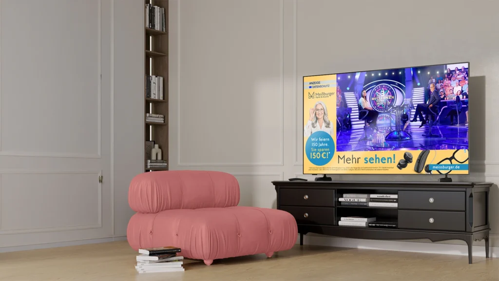 Modernes Wohnzimmer mit rosa Sessel und Sideboard, auf dem ein Fernseher steht. Auf dem TV läuft eine Quizshow mit eingeblendeter Werbung eines Hörgeräteanbieters auf der linken Seite.